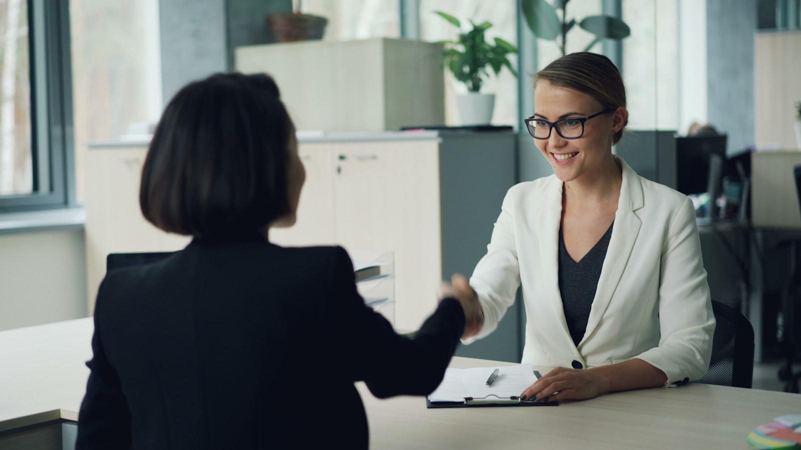 A recruiter welcomes a candidate with a handshake across a desk